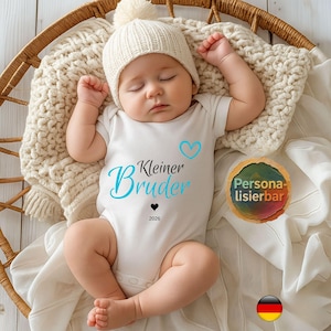May include: A sleeping baby wearing a white onesie with the words "Kleiner Bruder" in blue and black, a blue heart, and the year 2026. The baby is also wearing a white knit cap with a pom-pom. The baby is lying on a cream-colored blanket.