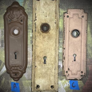 May include: Three antique door hardware pieces in varying shapes and colors. The pieces include a brown, ornate plate, a long, off-white plate, and a pinkish-beige plate. Each plate has a keyhole and a circular opening.