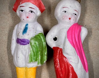 Puede incluir: Dos figuritas de porcelana vintage, una con un sombrero rojo y ropa verde y morada, la otra con una túnica rosa y roja con una corona dorada.