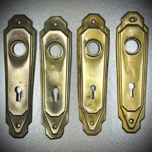 May include: Four vintage brass door plates with keyholes and circular handle openings. The plates have a rectangular shape with decorative edges and a tarnished, aged appearance. The plates are arranged in a row.