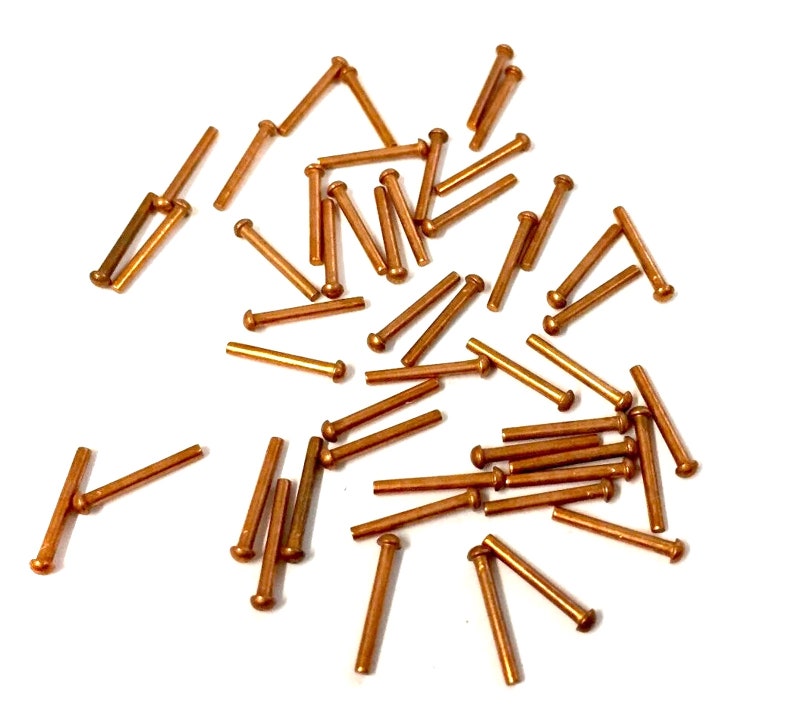 May include: A large quantity of copper-colored metal rivets, scattered on a white surface. The rivets are small and cylindrical with a rounded head on one end.