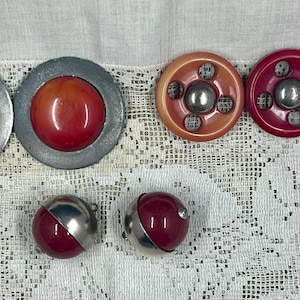 May include: Collection of vintage buttons in various styles and colors. Includes two large, round buttons with red centers and silver rims, three smaller buttons with silver accents, and two spherical buttons with silver and red sections.