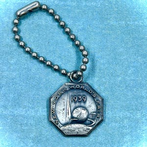 May include: Silver key chain with a round, octagonal charm featuring the text "New York World's Fair 1939". The charm depicts a stylized image of the Trylon and Perisphere, iconic structures from the 1939 World's Fair.