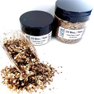 May include: Two clear plastic jars with black lids containing copper-colored mica flakes, labeled EK Mica / Flake Small Flake / Copper with Net Wt. 1oz and 2oz. A clear plastic scoop holds a portion of the metallic flakes, spilling onto the surface.
