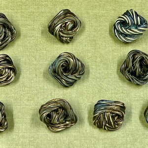Può includere: Vista dall'alto di undici bottoni decorativi quadrati. Ogni bottone è realizzato con filo ritorto, con tonalità argento, oro e verde-blu. I bottoni sono disposti su una superficie verde chiaro, creando un effetto strutturato.