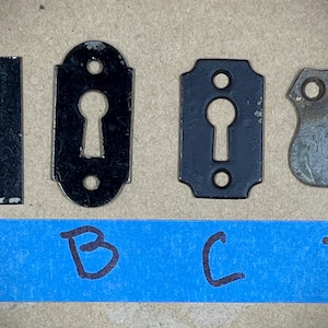 May include: Four different keyhole escutcheons, labelled A, B, C, and D. The escutcheons are made of metal and have a black finish. The escutcheons are in the shape of a rectangle, oval, rectangle, and shield.