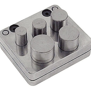 May include: A silver metal disc cutter tool with five cylindrical posts of varying heights. The tool is square with rounded corners and has two small holes and a screw. The tool is designed for jewellery making.