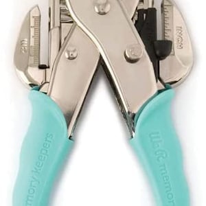 May include: A silver and teal We R Memory Keepers crop-a-dile tool. The tool has a silver metal body with teal handles. The handles are curved for grip and have the words "We R Memory Keepers" printed on them. The tool has a ruler on the side.