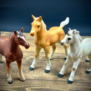 Puede incluir: Tres figuritas de caballos de porcelana. Una es marrón, otra es blanca y otra es marrón claro. Los caballos están de pie sobre una superficie de madera.