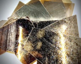 May include: A stack of thin, translucent, gold-toned sheets with a slightly iridescent sheen. The sheets are stacked in a random, overlapping pattern, creating a textured, abstract design.