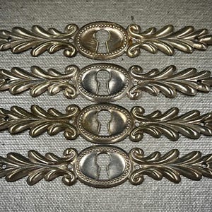 Puede incluir: Cuatro cubiertas de cerradura antiguas de latón con un diseño de hojas ornamentado. Cada cubierta tiene un ojo de cerradura ovalado y está rodeada por un borde con cuentas. Las cubiertas son simétricas.