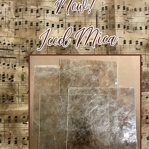 May include: Four sheets of iridescent mica, a type of mineral, in various sizes. The mica is translucent and has a silvery-gold sheen. The mica is arranged in a rectangular shape on a brown background.