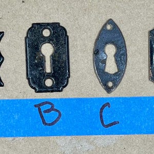 以下が含まれることがあります： A、B、C、D とラベル付けされた 4 つの黒い金属製の鍵穴エスカッション。エスカッションは、形とサイズが異なります。