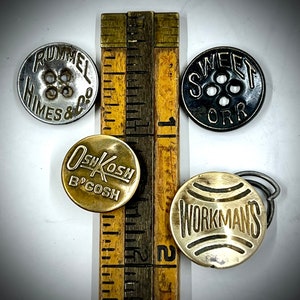 May include: Assortment of six vintage metal buttons with different designs and text. The buttons are round and have a shiny finish. The text on the buttons includes "Rummel Himes & Co", "Sweet Orr", "Oshkosh B'Gosh", and "Workmans".