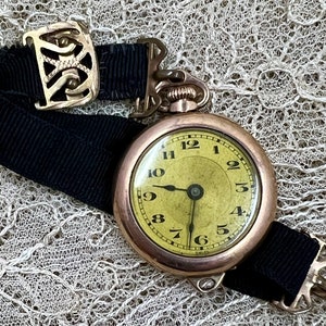 Könnte beinhalten: Eine goldene Taschenuhr mit einem schwarzen Band und einem gelben Zifferblatt. Die Uhr hat römische Ziffern für die Stunden und schwarze Zeiger. Die Uhr liegt auf einem weißen Spitzenhintergrund.