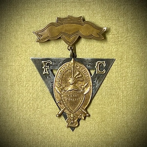 May include: A gold and silver triangular pin with the words "Knights of Pythias" and the letters "F" and "C" on it. The pin features a knight in armor with a sword and shield.