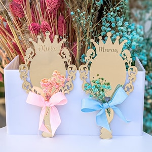 May include: Two gold-colored, crown-shaped place cards with the names "Olivia" and "Marcus" inscribed. Each card features an oval center, decorative filigree, and a small bouquet of flowers tied with a ribbon. One has pink flowers and ribbon, the other blue.
