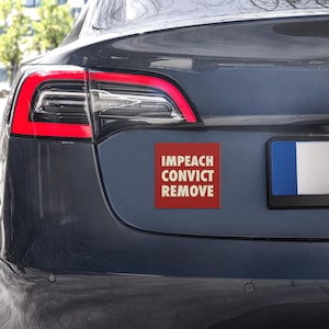 May include: A red square magnet with the words "IMPEACH CONVICT REMOVE" in white text is affixed to the rear of a dark gray car. The car's taillight is visible, with a red outline. The license plate is partially visible.