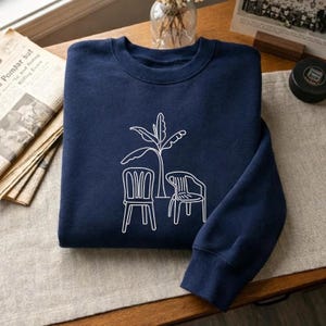 Puede incluir: Sudadera azul marino con un dibujo lineal blanco de una planta y dos sillas. La sudadera está doblada sobre una superficie de madera. El diseño es minimalista y adecuado para el uso diario.