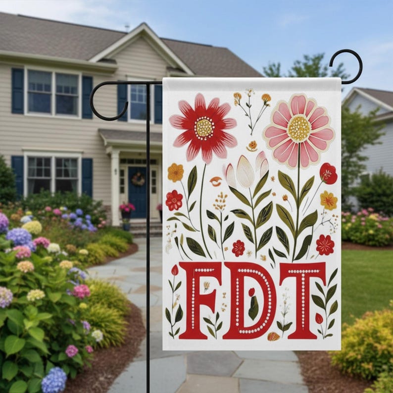May include: A white garden flag featuring a floral design with red and pink flowers, green leaves, and the letters "FDT" in red. The flag is displayed in a garden setting with a house in the background.