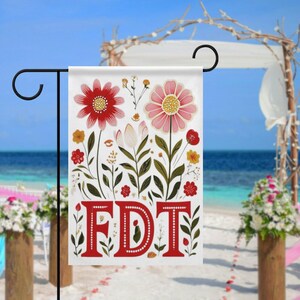 May include: A white garden flag with a floral design featuring red and pink flowers, green leaves, and the red letters "FDT". The flag is displayed on a black metal stand, with a beach and ocean background.