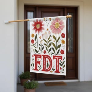May include: A white garden flag with a floral design and the red letters "FDT". The flag features red and pink flowers, green leaves, and yellow accents. The flag is hanging on a wooden pole.