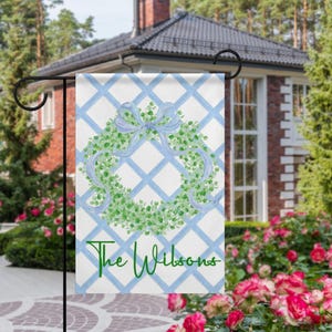 May include: A garden flag with a green wreath and blue bow design on a white and blue lattice background. The flag reads "The Wilsons" in green script. The flag is displayed outside a house, with a garden in the foreground.