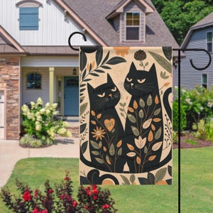 Puede incluir: Una bandera de jardín con dos gatos negros rodeados de diseños florales y de hojas. El diseño utiliza tonos beige, naranja, verde y marrón. La bandera está colgada frente a una casa, agregando un toque decorativo al jardín.