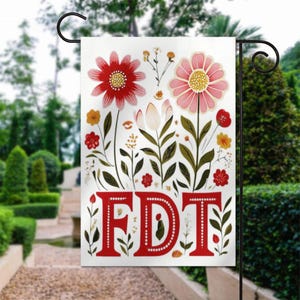 May include: A white garden flag featuring a floral design with red and pink flowers, green leaves, and the red letters "FDT". The flag is displayed on a black metal stand in a garden setting.