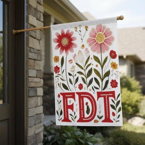 May include: A white garden flag with a floral design featuring red and pink flowers, green leaves, and the letters "FDT" in red. The flag is hanging on a wooden pole outside a house.