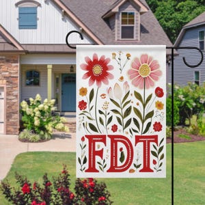 May include: A white garden flag with a floral design and the letters "FDT" in red. The flag features red and pink flowers, green leaves, and yellow accents. The flag is hanging on a black metal stand in front of a house.