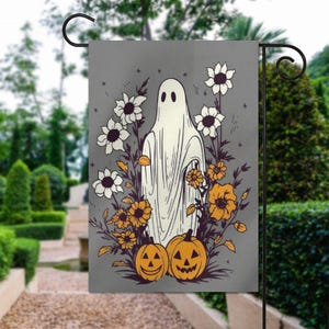 May include: A Halloween garden flag featuring a white ghost figure surrounded by white and orange flowers, and two jack-o'-lanterns. The background is a muted gray, and the flag is displayed on a black metal stand.