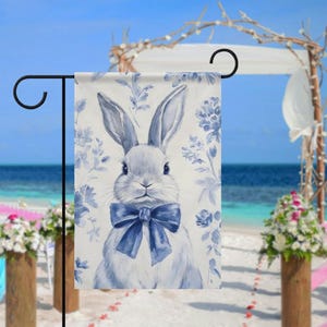 May include: A garden flag with a watercolour illustration of a rabbit wearing a blue bow tie. The rabbit is set against a backdrop of blue floral patterns. The flag is displayed on a black metal stand, with a beach and ocean in the background.