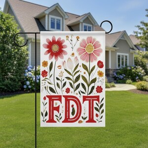 May include: A white garden flag with a floral design and the letters "FDT" in red. The flag features red and pink flowers, green leaves, and other floral elements. The flag is displayed on a black metal stand in a yard.