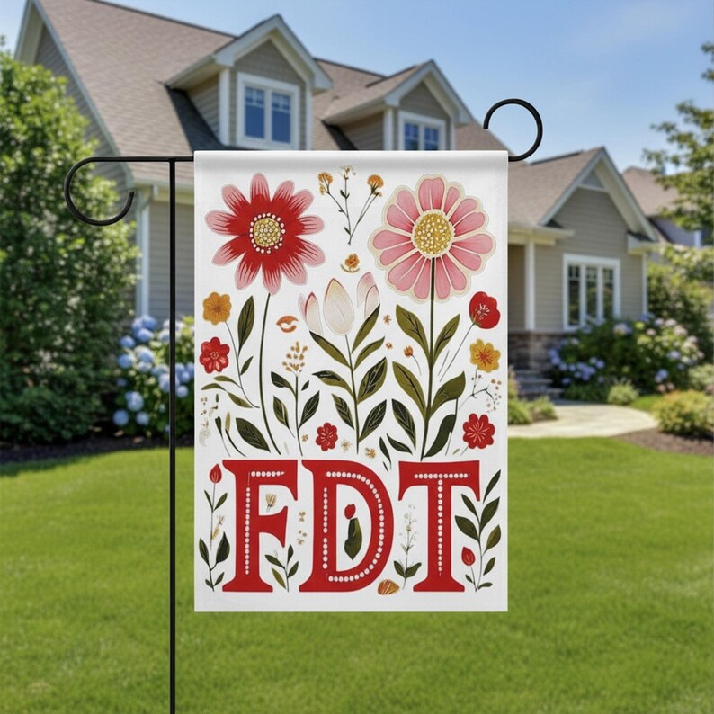 May include: A white garden flag with a floral design and the letters "FDT" in red. The flag features red and pink flowers, green leaves, and other floral elements. The flag is displayed on a black metal stand in a yard.