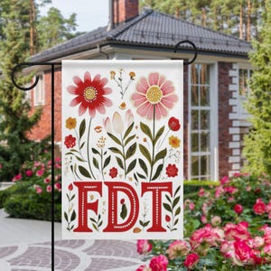 May include: A white garden flag featuring a floral design with red and pink flowers, green leaves, and the letters "FDT" in red. The flag is displayed on a black metal stand in a garden setting.