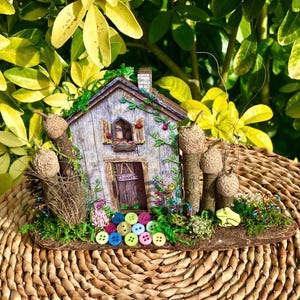May include: A miniature house sculpture featuring a weathered wood-look facade, a small door with a sign, and shuttered windows. The structure is decorated with acorn-topped posts, green moss, colourful plastic buttons, and a small yellow frog figure.