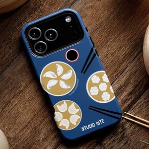 Könnte beinhalten: Eine blaue Handyhülle mit einem Knödel-Design und dem Text "STUDIO BITE". Die Hülle hat ein kreisförmiges Design mit Knödel-Illustrationen in Gold und Weiß. Die Handyhülle liegt auf einer Holzoberfläche mit Knödeln und Tee.