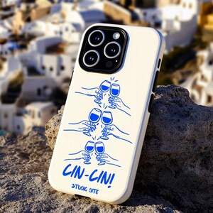 Könnte beinhalten: Cremefarbene Handyhülle mit einer blauen Illustration von Händen, die auf Weingläser anstoßen. Der Text "CIN-CIN! STUDIO BITE" ist blau gedruckt. Die Hülle liegt auf einem Felsen mit einem Hintergrund aus weißen Gebäuden und dem Meer.