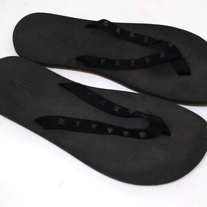 May include: A pair of black leather flip-flops with a black strap and a studded design. The flip-flops have the word "VALENTINO" printed on the strap.