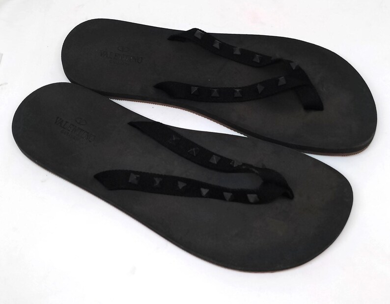 May include: A pair of black leather flip-flops with a black strap and a studded design. The flip-flops have the word "VALENTINO" printed on the strap.