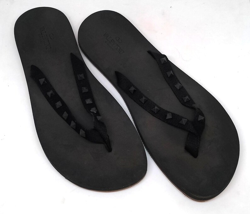 May include: A pair of black leather flip flops with a studded strap. The strap has the word "VALENTINO" printed on it.