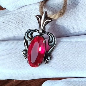 May include: A silver pendant with a large, oval, ruby-red gemstone. The pendant has an ornate, swirling design around the stone and is suspended from a natural fiber cord. The gemstone is approximately 1 inch in length.