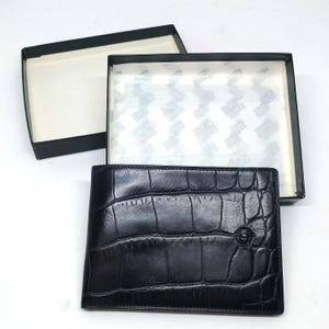 Może przedstawiać: Czarny skórzany portfel o fakturze skóry krokodyla, prezentowany w czarnym pudełku z białym wnętrzem. Portfel posiada mały, okrągły emblemat z przodu.