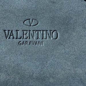 May include: A close-up of a dark blue suede surface with the embossed text "VALENTINO GARAVANI" and a stylized "V" logo.
