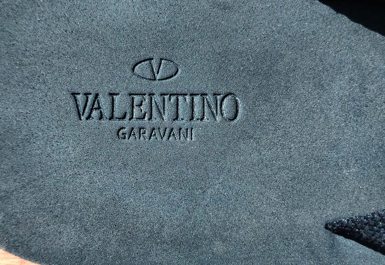 May include: A close-up of a dark blue suede surface with the embossed text "VALENTINO GARAVANI" and a stylized "V" logo.