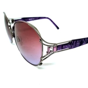 Puede incluir: Gafas de sol ovaladas con lentes rosas degradadas y montura de metal plateado. Las patillas son de color púrpura texturizado y el puente es de color púrpura a juego. Las gafas tienen un diseño moderno y elegante.