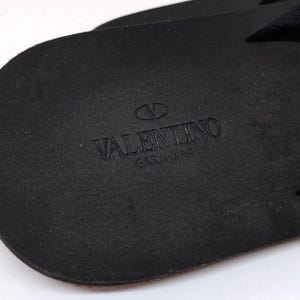 May include: Close-up of the black leather sole of a sandal with the Valentino Garavani logo embossed in the center.