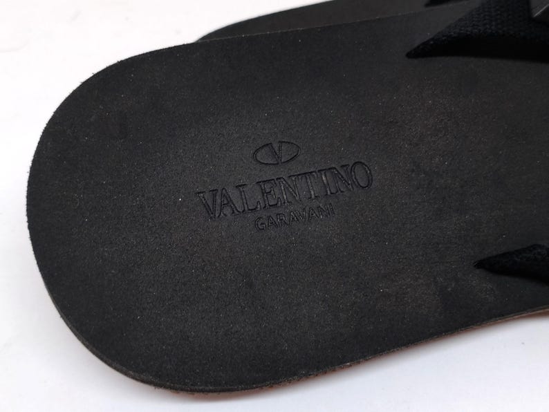 May include: Close-up of the black leather sole of a sandal with the Valentino Garavani logo embossed in the center.