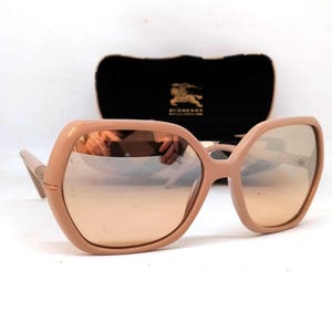 May include: Oversized, peach-colored sunglasses with a hexagonal frame and mirrored lenses. The sunglasses are set against a black case with a gold logo. The arms of the sunglasses are a lighter shade of peach.